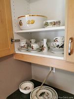 Kitchen cabinet shelf with large bowl and cups, saucers with fruit and vegetable designs, showing arrangement and style.