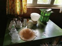 Multiple vintage light covers and fixtures spread on green table under window, showing varied frosted and clear patterned glass shades with multiple styles.