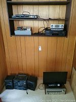 Photo showing shelf with Samsung DVD player, Panasonic clock radio, cassette tapes, Electrohome VHS player, Pioneer stereo receiver. Below shelf is Panasonic portable stereo with two speakers and RCA monitor with remote on a separate device.