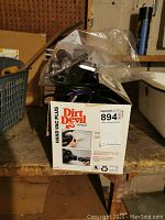 Photo showing Dirt Devil handheld vacuum in its original box with attachments packed in plastic. Box is placed on a wooden surface with background items showing.