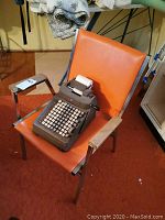 Front view of vintage orange office chair with chrome armrests, with manual adding machine placed on the seat.
