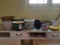 Wide view showing all 10 planters in a row on a shelf beneath a window, revealing their variety in color, size, and material.