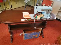 Wide-angle view of a vintage Singer sewing machine mounted in a wooden sewing table, showing the table top and some accessories below.
