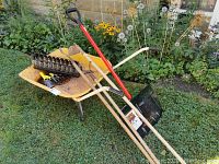 Photo shows a yellow wheelbarrow with dirt, a red-handle shovel, two wooden-handle garden tools (rake and hoe), a black metal rake head, and a decorative black metal tea light holder.
