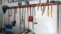 Wide shot of various hanging gardening tools including rakes, shovels, pruners, and other implements attached to a wooden rack.