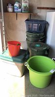 Photo shows green plastic laundry bin with white rope handles in front, green garden bins with lids behind it, dark grey laundry basket, white and green storage tote, orange small bucket, and cleaning supplies on shelf above.