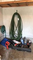 Full view of lot in garage showing large green garden hose on wall hook, smaller coiled green hose, plastic watering cans, and Black and Decker leaf blower with attachments on floor.