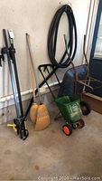 Photo of metal roof rack, two straw brooms, yellow dustpan, Scotts Easy Green II seeder, and old metal wheelbarrow with rust and corrosion