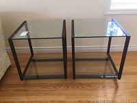 Pair of matching metal-framed side tables with glass tops and lower shelves placed on wood floor next to couch.