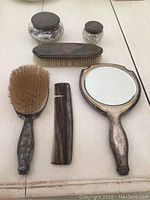 Photo of all six vanity items displayed on a flat surface showing handheld mirror, hairbrush, comb, clothes brush, and two cut glass jars with silver lids.