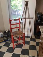 Photo showing the wooden painter's easel and red painted kitchen chair with rush seat, positioned on a checkered floor near a window.