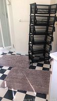 Photo shows two taupe colored rectangular bath mats placed on checkered floor, with six black stacking plastic storage bins arranged behind them.