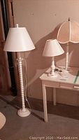 Set of three white lamps: tall painted wood floor lamp with spindle detail, and two vintage style table lamps with fabric shades shown on and next to white table