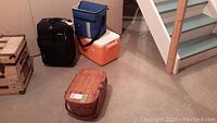 View of all four items: vintage picnic basket, black Delsey suitcase, Coleman cooler, and blue California Innovations cooler bag arranged on concrete floor near stairs.
