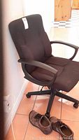 Side angle of the brown fabric office chair showing padded armrest and five-point rolling base, with a shoe beside it for scale.