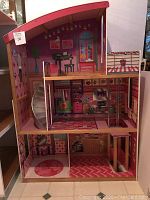 Front and side angled view of wooden dollhouse showing three floors, silver elevator winding upwards, top balcony with black piano, and pink decorative interiors.