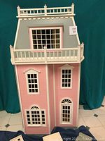 Front view of the pink wooden doll house showing three levels with multiple windows and balcony.