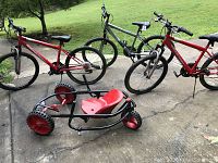 Front view of two matching red Roadmaster boys bikes, one silver Roadmaster mountain bike, and a black-framed kid's Y-bike with red seat on concrete driveway with grass in the background.