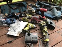 Overview of tools and accessories laid out on wooden deck showing tool bags, power saw, drills, sanders, charger and angle grinder.
