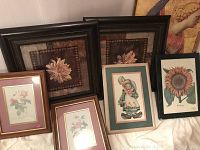 Photo showing five framed artworks arranged leaning together. Includes two large flower paintings with dark borders, two botanical rose prints with maroon and wooden frames, and one illustration of a child-like character in green frame.