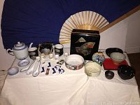 Photo of vintage Asian collection including teapot, various bowls, porcelain spoons, cups, decorative hand-painted case, and folding fan.