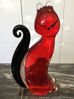 Side view of vintage Murano glass cat sculpture with red body, black tail and clear glass base shown on marble surface against white background.