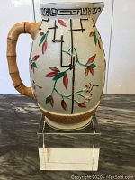Full view of the jug showcasing Japanese styled painted bamboo and leaves, bamboo-style handle, standing on a clear acrylic block.