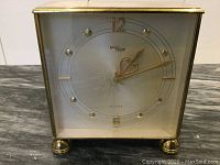 Frontal clear image of the IMHOF clock face showing frosted gold and white dial, golden numerals, and brass case.