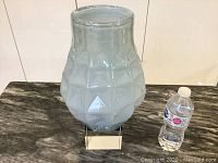 Large modern iridescent glass vase on a reflective stand next to a water bottle for scale.