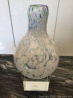 Front angle showing the rounded bulbous body with white speckled pattern and hints of blue and yellow. The vase stands on a clear acrylic block.