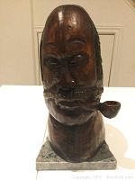Front view of primitive wooden carving bust of man smoking a pipe on a square base, showing facial details and wood finish.