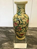 Full front view of the vintage Asian ceramic vase on a clear acrylic stand showing the painted branches, leaves, flowers, and fruits against a cream-yellow background.