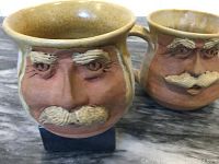 Two handmade pottery pieces, a cream jug and sugar pot, with sculpted faces. Both have mustaches and eyebrows, showing artisan craftsmanship.