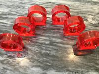 Six oval red Lucite napkin rings arranged in two groups of three on a marble surface, showing their semi-transparent ruby red color and flat base.