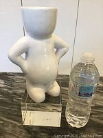 Front view of white ceramic person vase with hands on hips, on a clear acrylic stand next to a water bottle for scale.