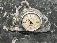 Front angle shot of Etain Zinn Seagull signed pewter clock on a dark marbled surface showing floral ornate design and clock face with Roman numerals.