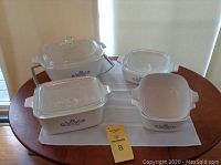 Set of 5 Corning Ware casserole dishes in white with blue cornflower design and clear glass lids, arranged on a round wood table with a white cloth underneath.