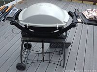 Front view of white Weber gas grill with lid closed on two-wheeled metal stand, skewers and utensils visible beside it.