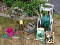 Full lot overview showing birdbath, pink and metal watering cans, garden hose reel, and sprinkler timer with fittings