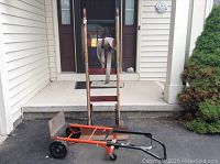 Front view of the two hand trucks/carts in front of a house entry. Brown hand truck with wooden handles stands behind the orange wheeled appliance cart.