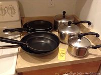 Set of 3 stainless steel Faberware pots with lids, and several black nonstick frying pans on a kitchen counter.