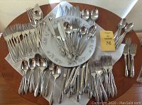 Wide view showing all flatware pieces arranged on wooden table around a floral plate and seller card. Includes forks, knives, spoons of differing designs.