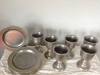 Full set of 6 pewter plates stacked with 6 pewter cups arranged behind them on a white cloth.