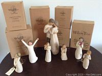 Overview of 8 Willow Tree resin figurines arranged on a table in front of original cardboard boxes. Figurines include angels, children, a couple embracing, and figures holding symbolic items.