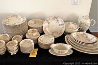 Full set displayed on table showing plates, teacups, saucers, serving platters, bowls, and coffee pot all with matching pink and cream floral pattern and scalloped gold edges. Three teacups show small chips.