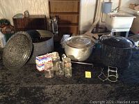 Wide view of enamelware pots, aluminum kettle, canning jars, wax, and jar grabbers on a black surface.