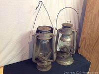 Two antique oil lanterns shown together on black surface against white backdrop, showing full front and side views.