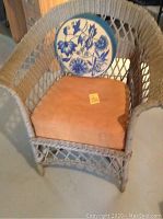Front angled view showing the wicker chair, orange seat cushion, and round blue floral needlepoint pillow inside the chair.