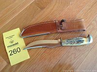 Fixed blade knife with bone handle and leather sheath laid on wooden floor next to auction lot number card.