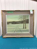 Front view of framed painting showing winter snow scene with tracks, two trees and distant hills in textured wood frame